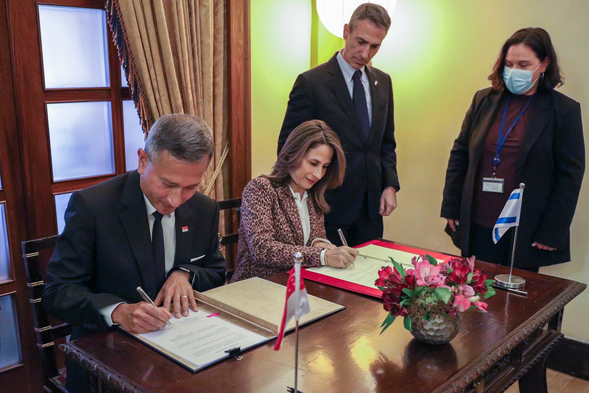Two people sign documents on a table with Singapore and Israel flags. Two people observe.